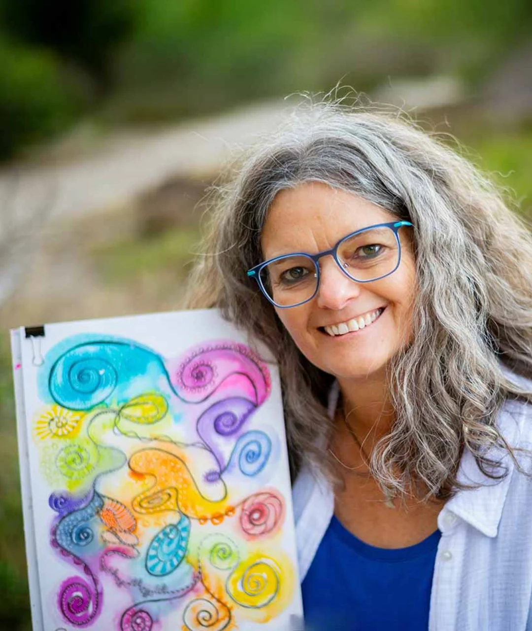 Christina lächelt im Grünen und hält eines ihrer bunten Kunstwerke in den Händen.
